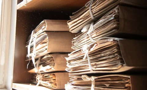 Two piles of dusty beige cardboard files containing paperwork sitting on a shelf with dust motes floating in a shaft of light