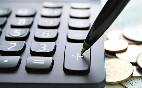 Calculator next to a pile of coins with a pen poised to press the plus key